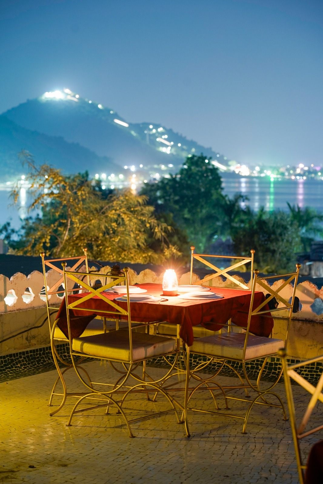 Rooftop dinner table lit at night at Vikram Garh Lakeview, with Fateh Sagar and the lit hillside behind